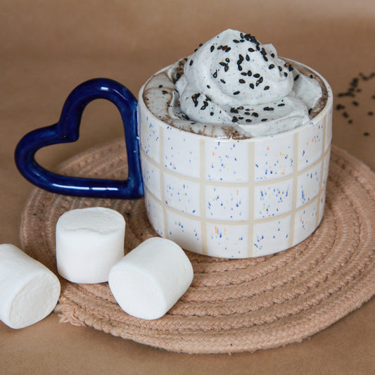 A black sesame hot chocolate topped with black sesame whipped cream in a tiled mug with heart-shaped handle, surrounded by marshmallows.