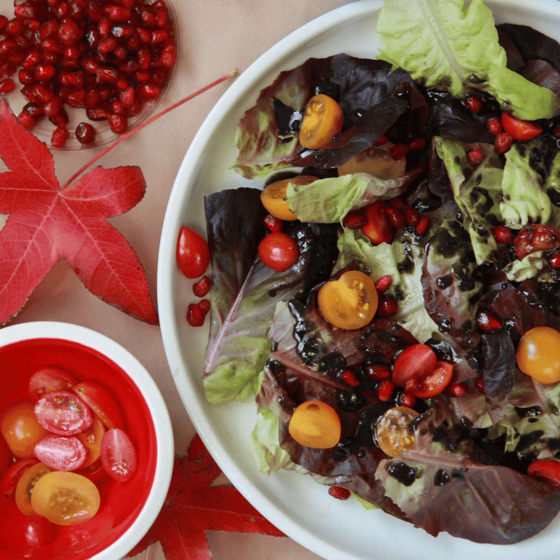 Colorful salad next to red maple leaf and two bowls of cut cherry tomatoes and pomegranate seeds.