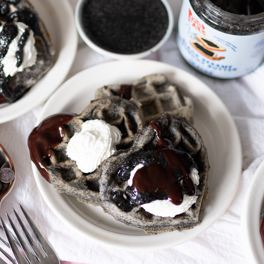 Heart-shaped black sesame waffles topped with whipped cream and chocolate sauce, made with Rooted Fare's Black Sesame Crunchy Butter