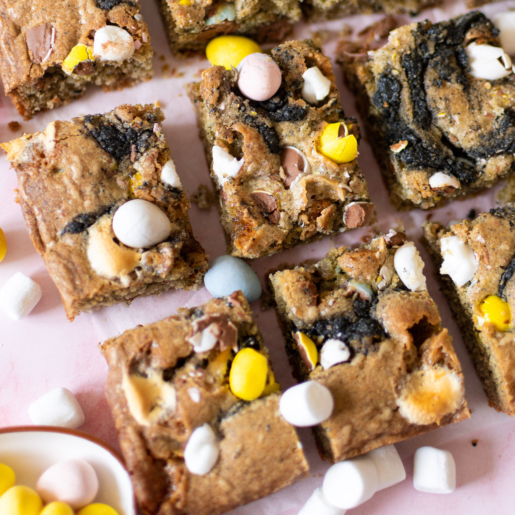 Close-up shot of cookie bars made with Rooted Fare's Black Sesame Crunchy Butter (swirled on top); colorful Cadbury eggs in pastel blue, pink, yellow, and beige; and tiny marshmallows. 