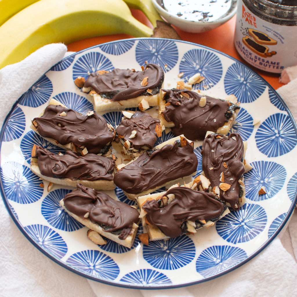 Bite-sized banana treats topped with black sesame spread, chocolate, and nuts, presented in a indigo and white plate.