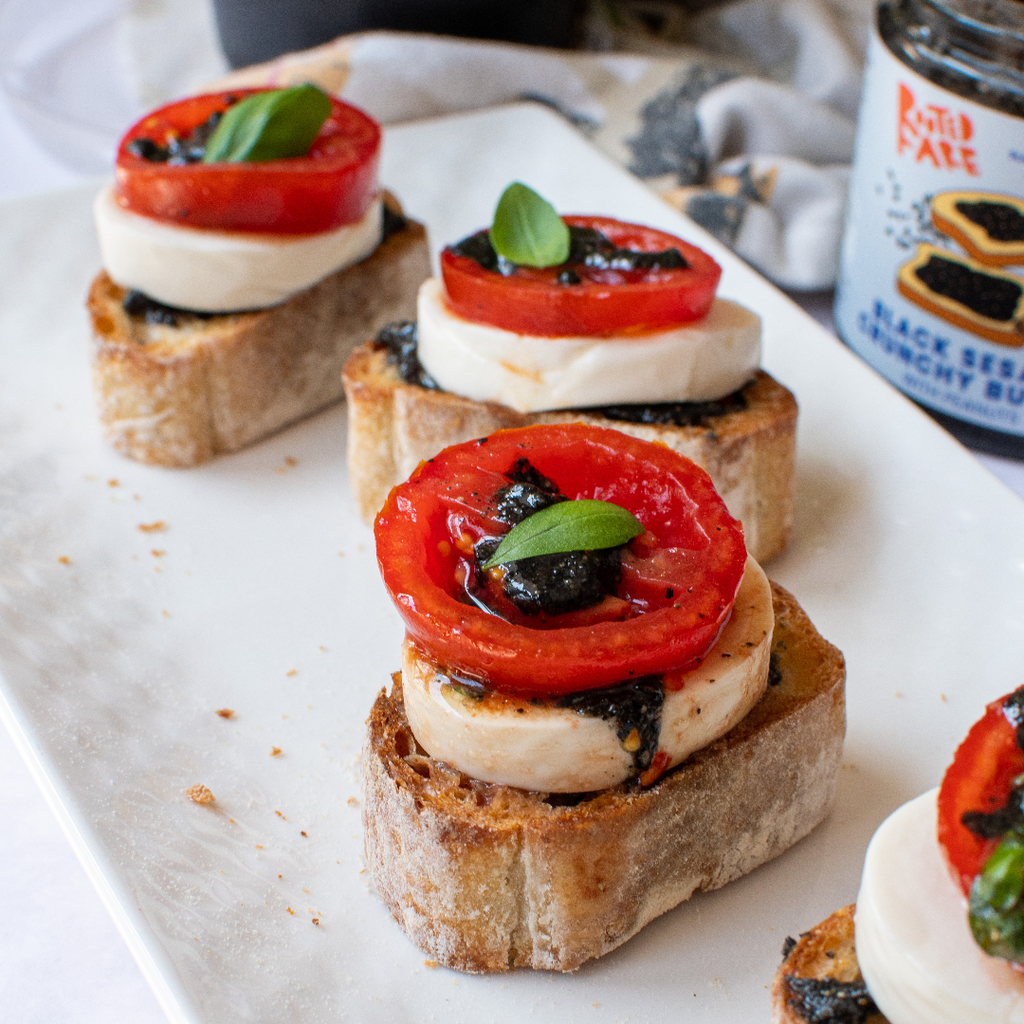 Four small pieces of toast with thick-sliced mozzarella, tomato, and a basil leaf, topped with Black Sesame Crunchy Butter dripping down the side. Plated on a white rectangular plate with a jar of Black Sesame Crunchy Butter in the background.