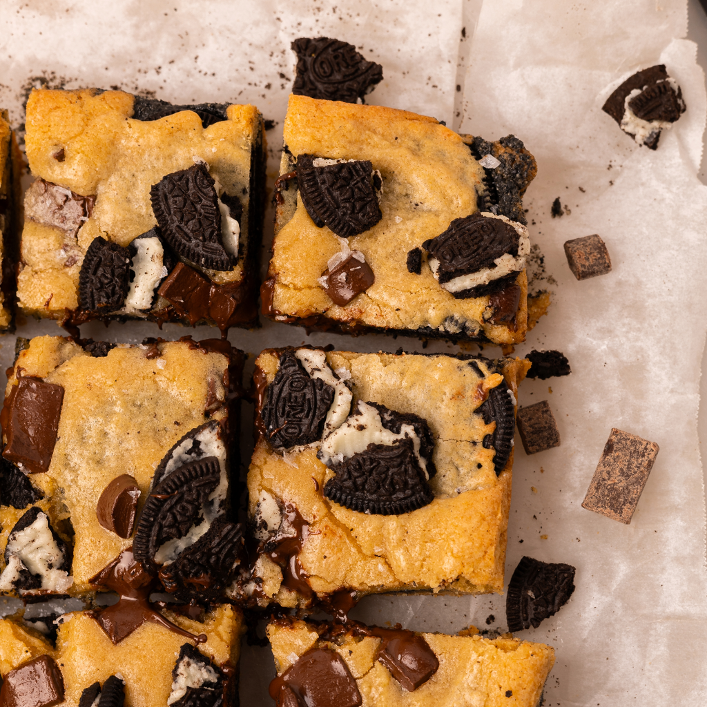 Squares of golden blondies with pieces of crushed oreos, gooey chocolate, and black sesame swirls, laid out on white parchment paper.