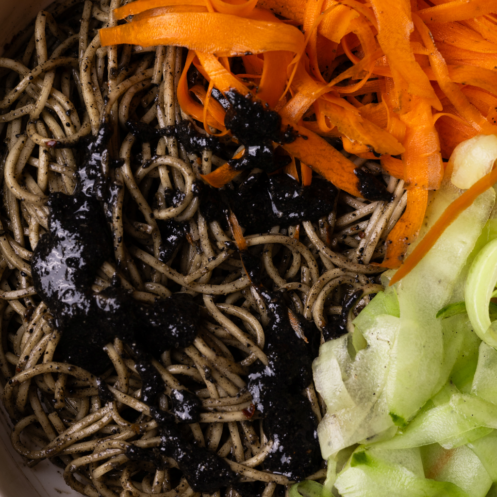 Close-up shot of noodles smothered in Crunchy Black Sesame Butter with slices of fresh cucumbers and carrots as garnish.