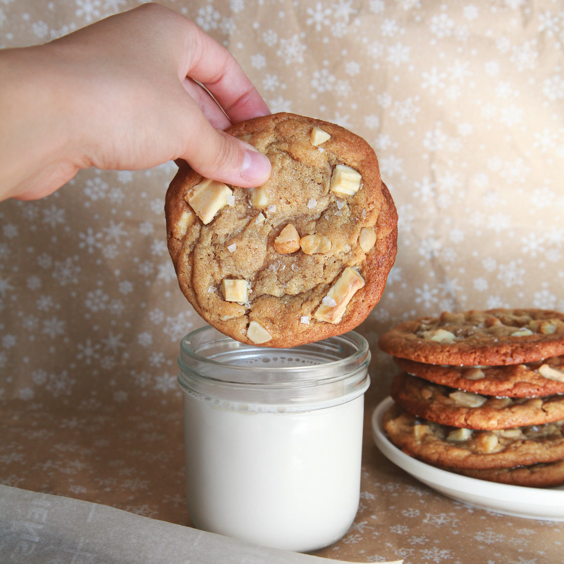 A large pineapple white chocolate macadamia cookie with golden brown edges, held above a glass of milk.