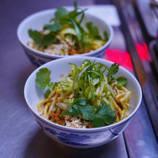 Two bowls of noodles topped with shredded cucumber, fresh cilantro, long strips of scallions, crushed peanuts, and brown sauce.