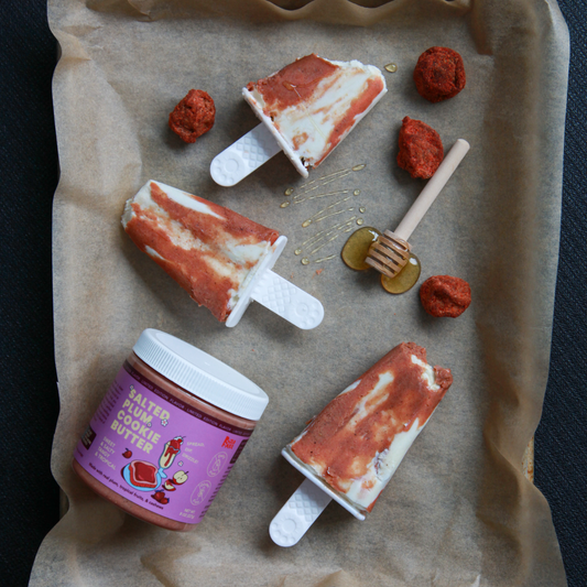 Sheet tray lined with parchment paper with three popsicles and ingredients on top. Popsicles have a creamy white base with burnt orange marbling made with Rooted Fare's Salted Plum Cookie Butter.