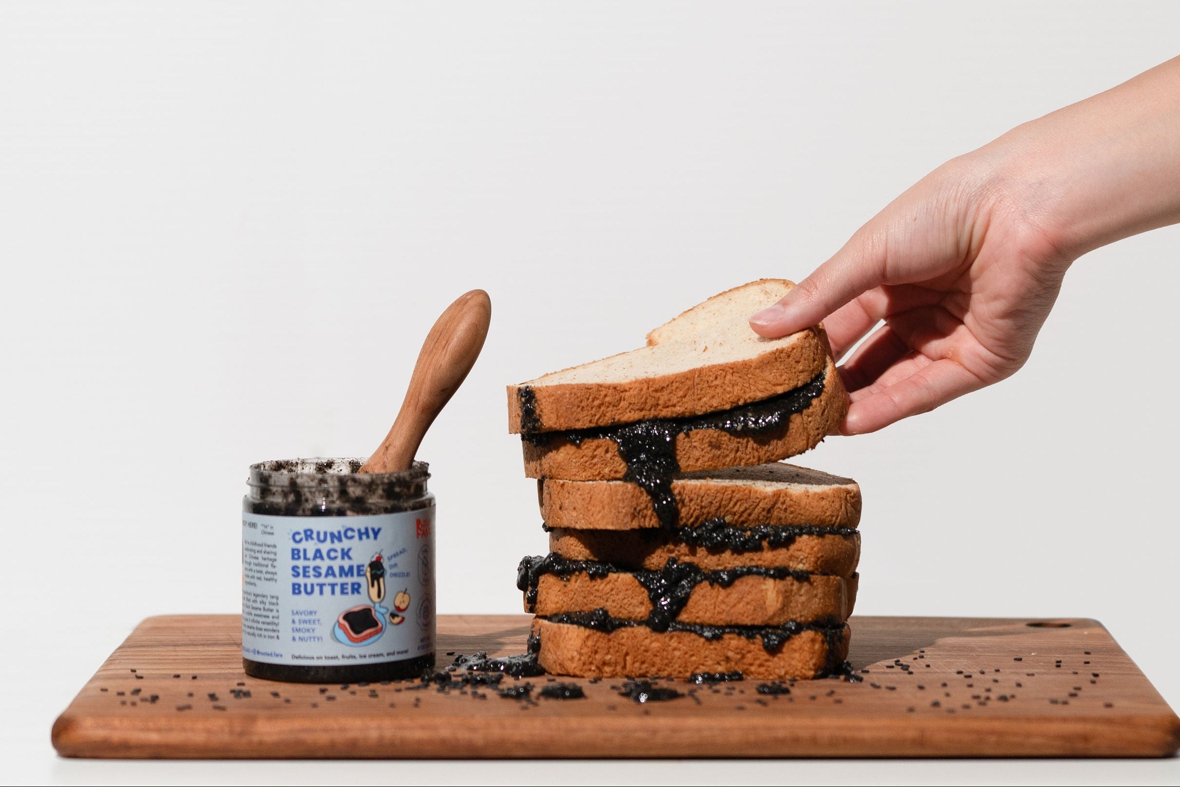 Hand reaching for a stack of toast with a can of jam on a wooden board against a white background